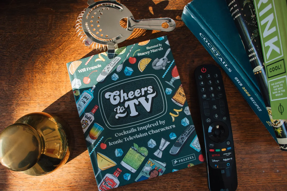 Cheers to TV by Will Francis, illustrated by Stacey Marsh, beside a television remote and cocktail equipment