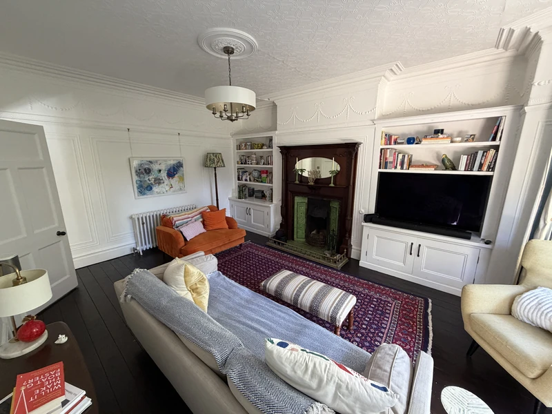 Original photograph of Will Francis’s sitting room, with an ornate white ceiling, wooden fireplace, green tiles, fitted shelves, orange loveseat and patterned rug