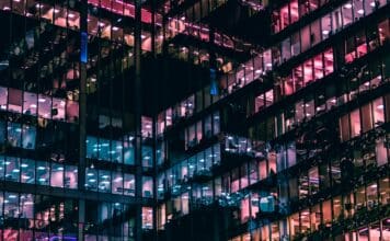 City office block windows at night with different colored lighting inside reflecting out off opposing glass walls to create an almost abstract geometric lattice of different colored lights.